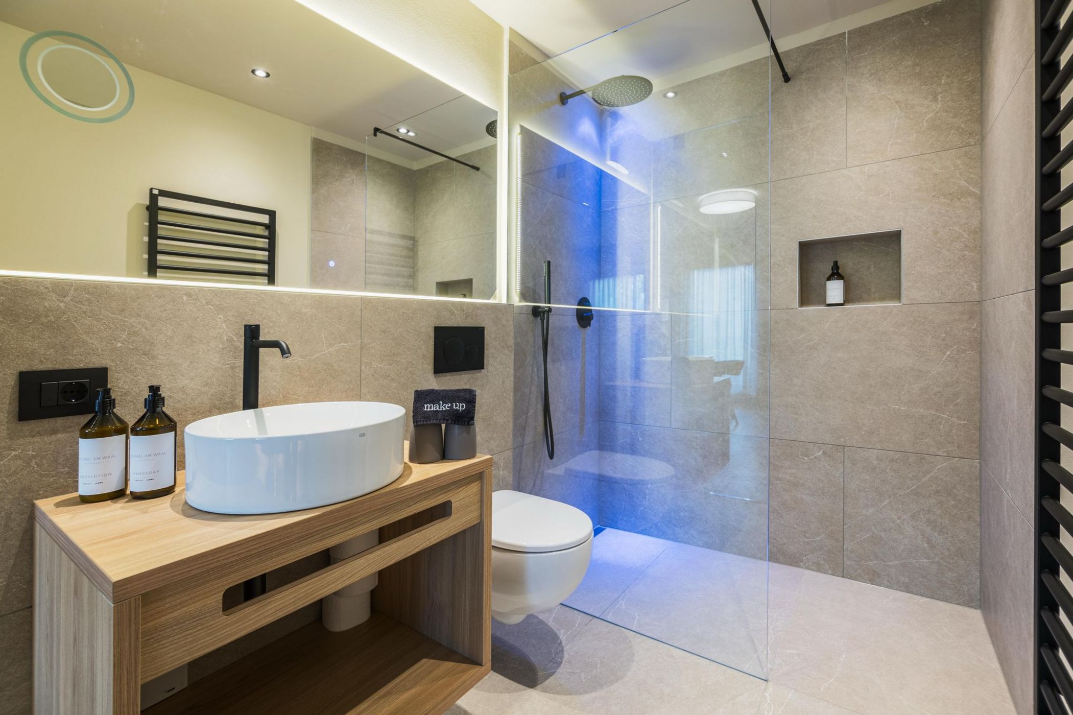 Bagno moderno con lavabo ovale bianco, specchio luminoso, doccia walk-in con parete di vetro e pareti in piastrelle beige.