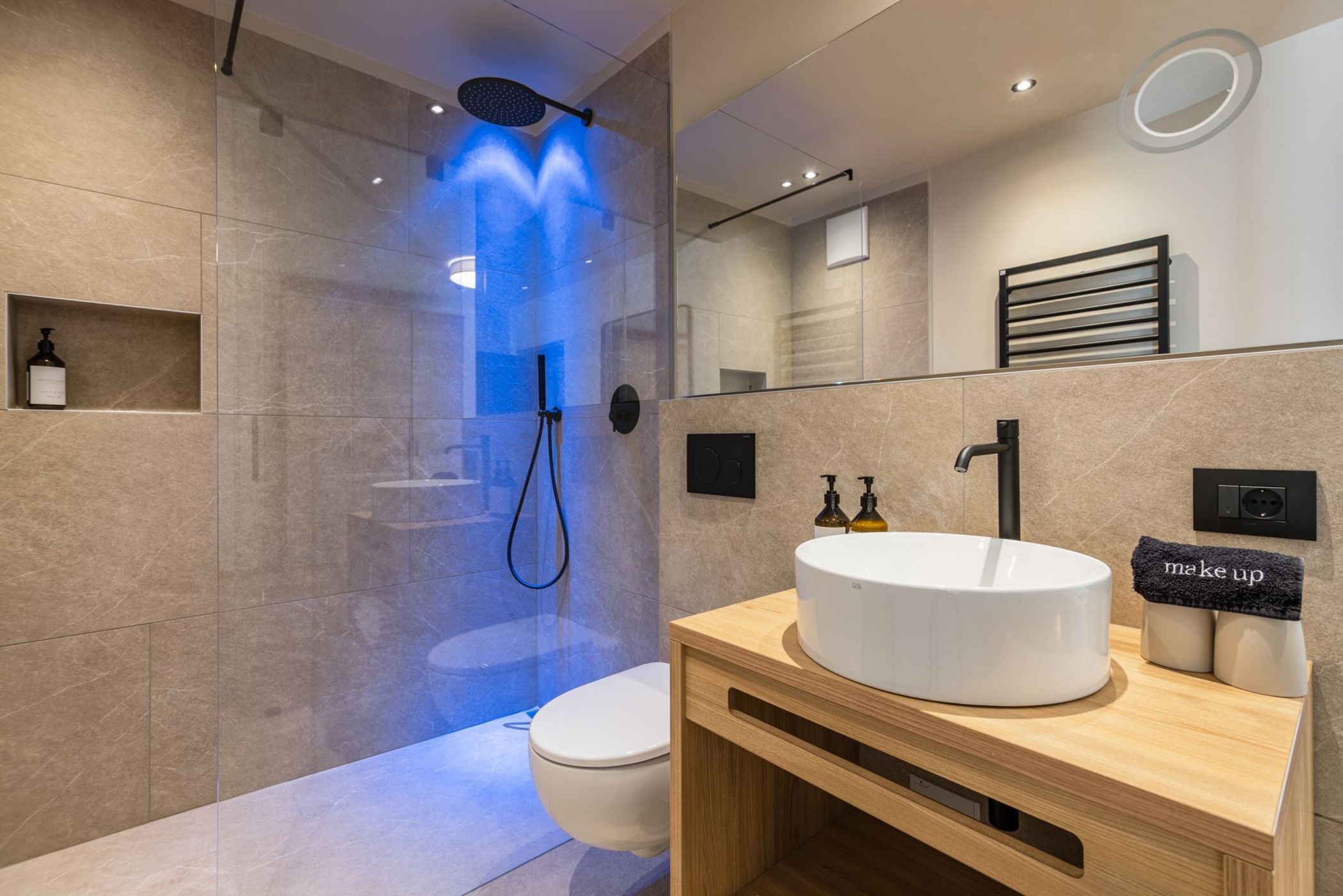 Bagno moderno con doccia a LED blu, lavabo bianco su mobile in legno e pareti beige.