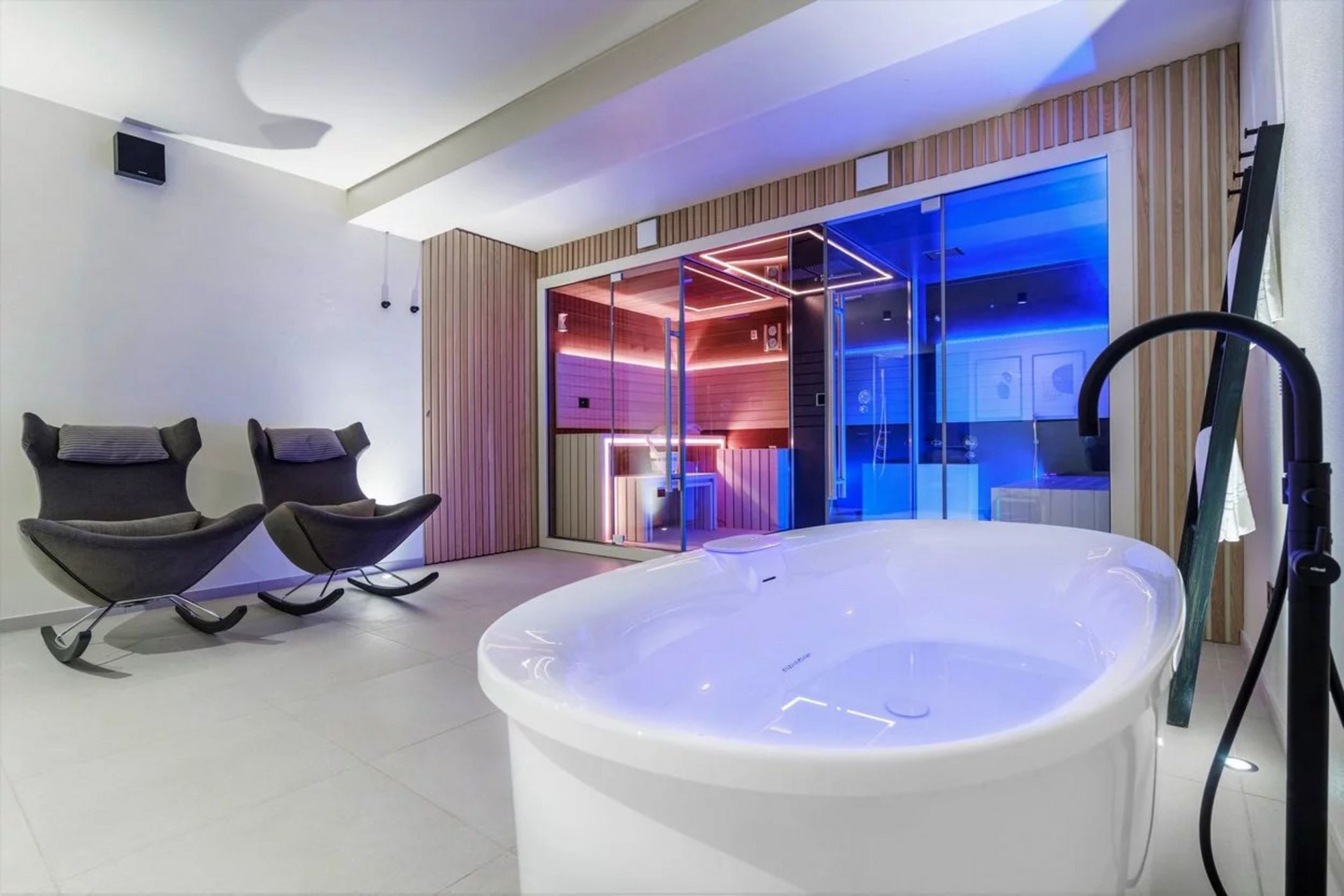 Sala spa moderna con vasca bianca, due poltrone grigie e sauna con luci colorate dietro una parete di vetro.