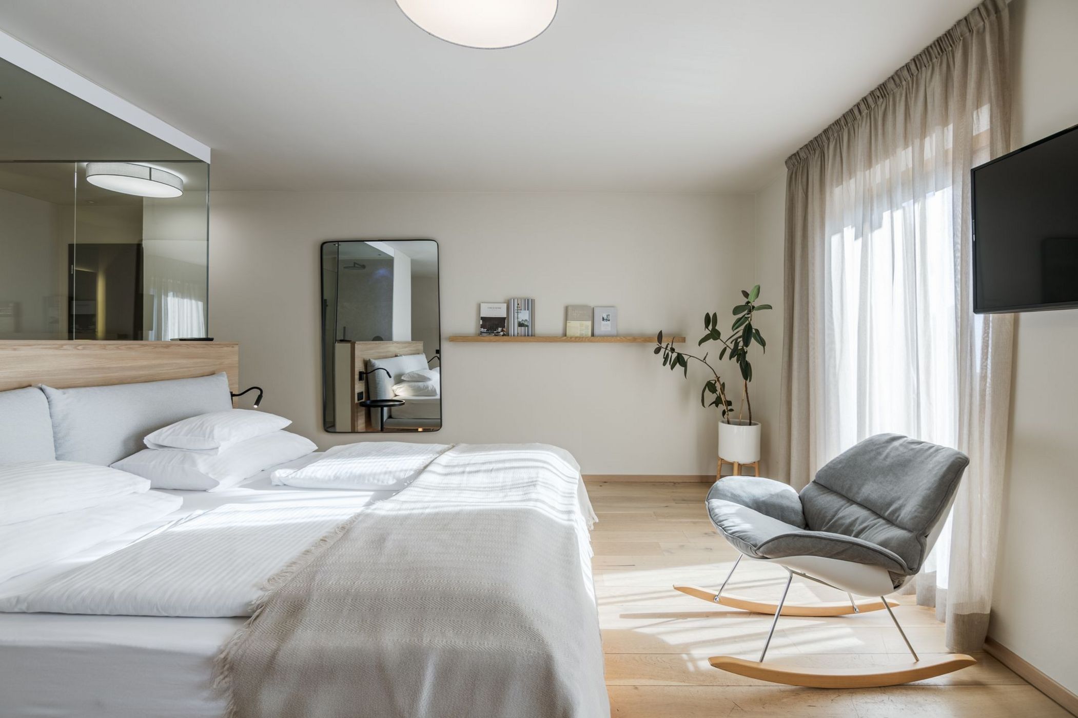 Camera da letto moderna con letto matrimoniale, sedia a dondolo grigia, specchio e pianta in vaso vicino alla finestra.