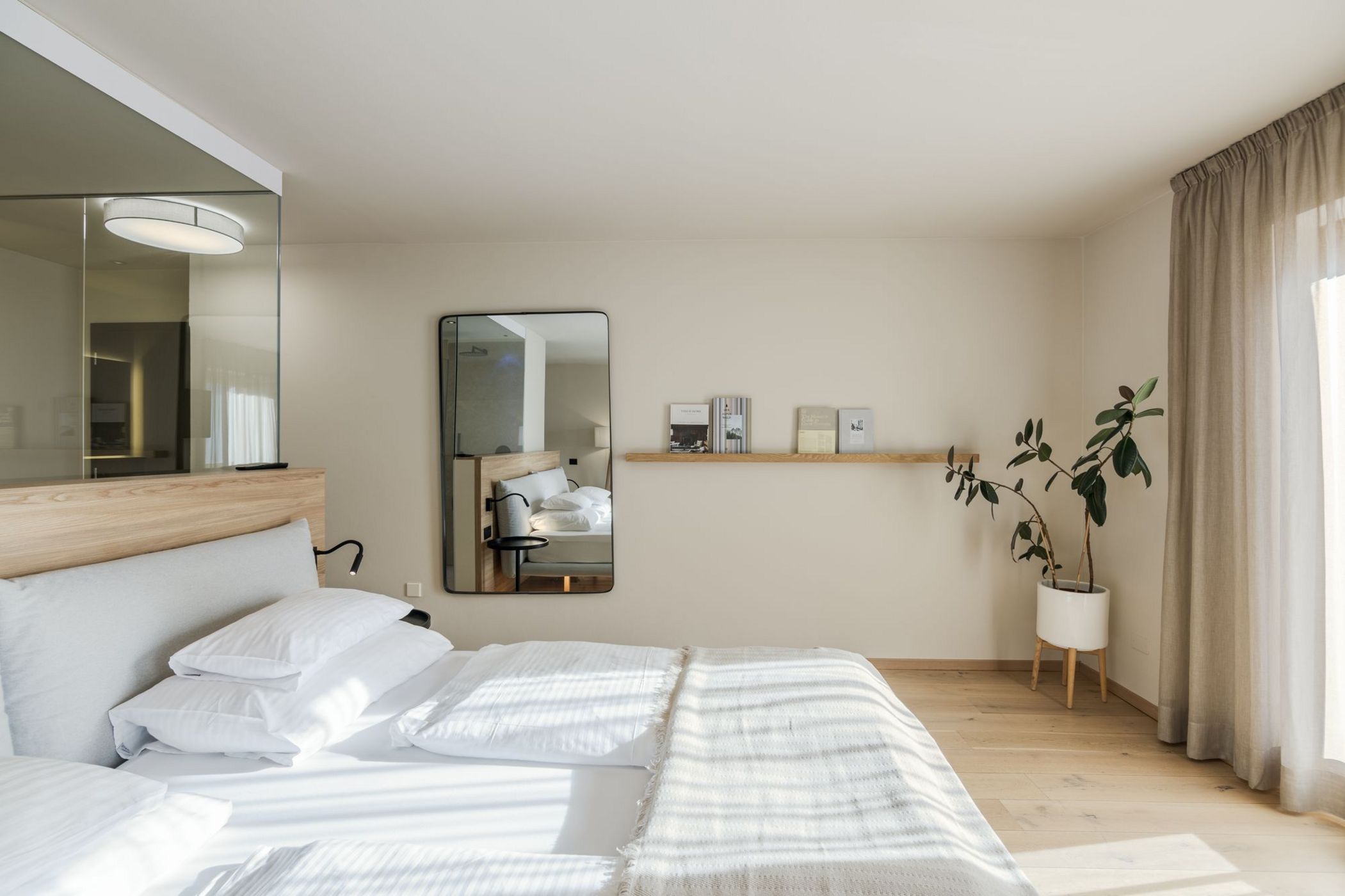 Camera da letto moderna con letto matrimoniale, specchio rettangolare, mensola con libri e pianta in vaso vicino a una finestra con tende.