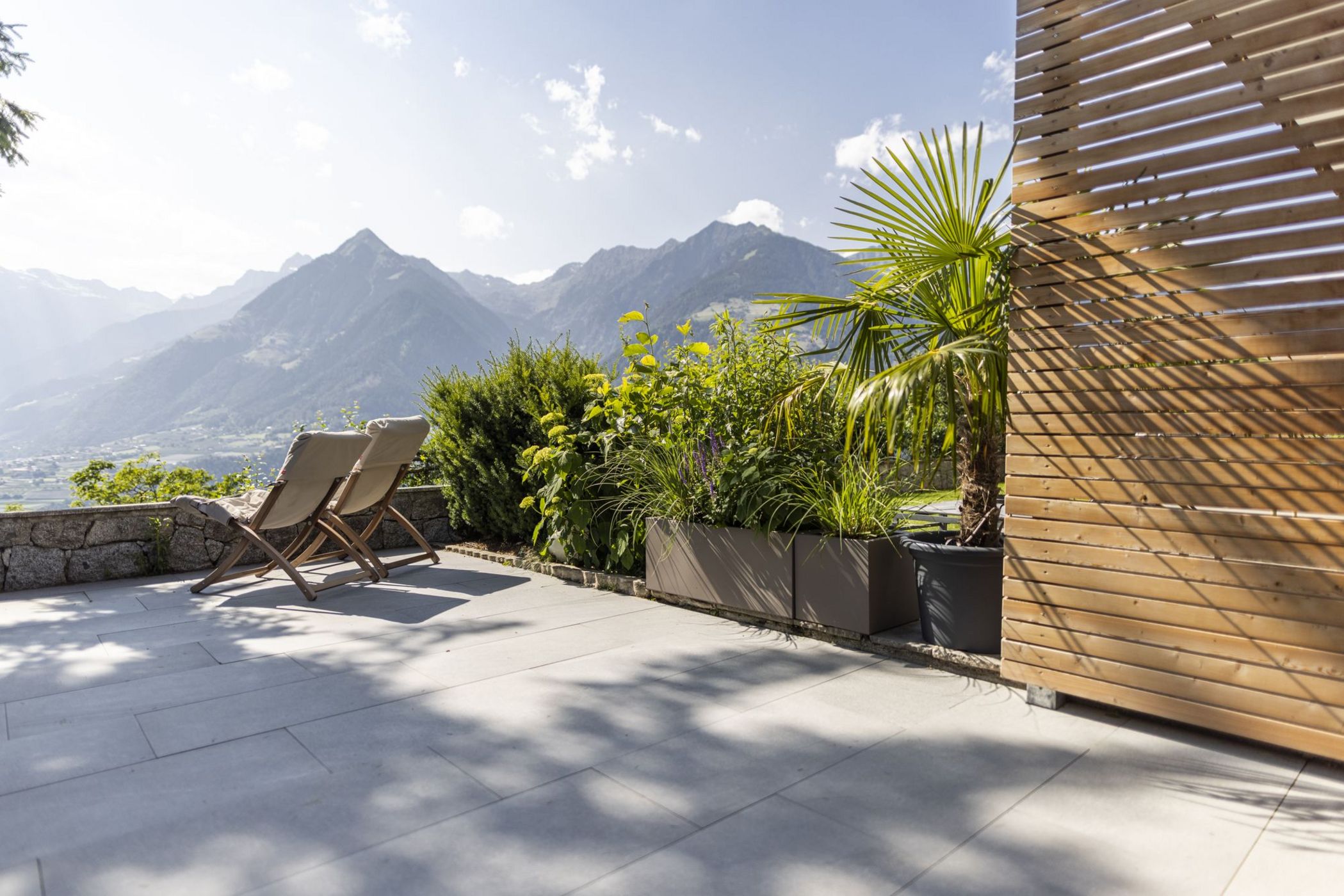 Terrazza con sedie a sdraio, piante e vista sulle montagne soleggiate.