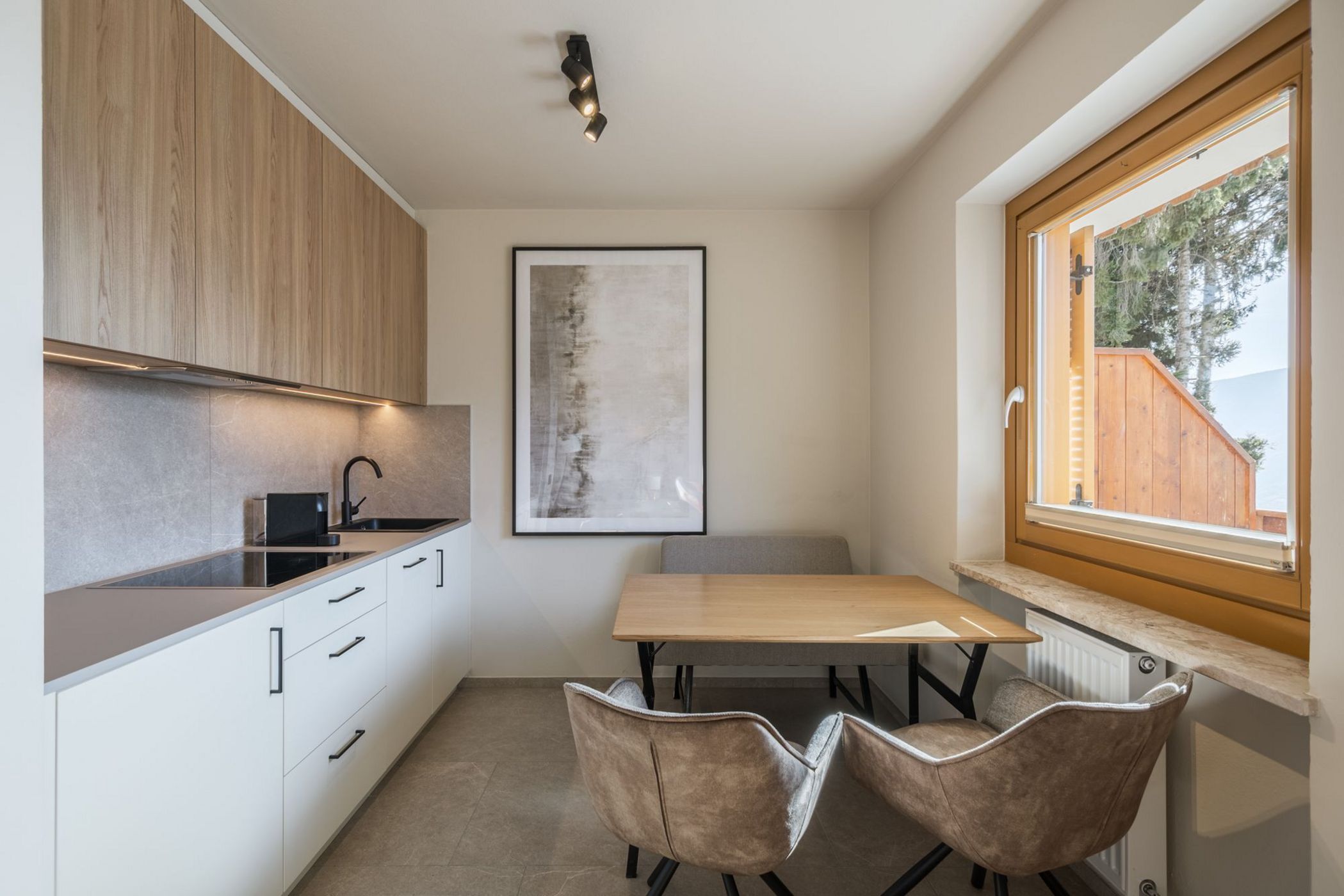 Cucina moderna con mobili in legno chiaro, tavolo di legno con due sedie imbottite e finestra grande con vista esterna.