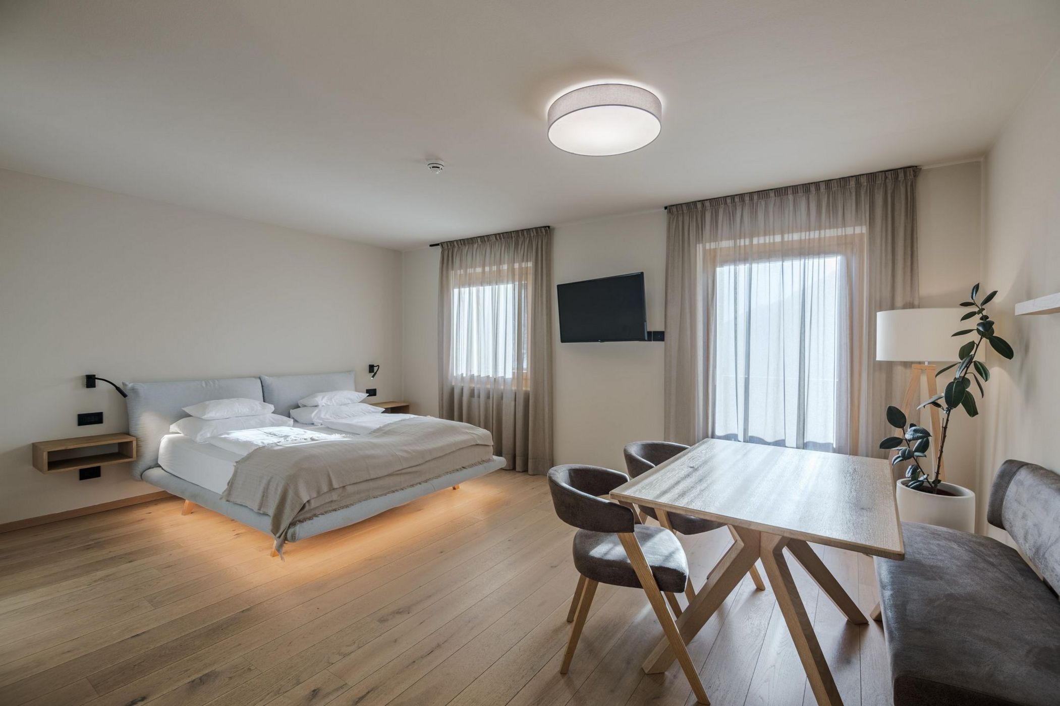 Camera da letto moderna con letto matrimoniale, tavolo in legno, sedie, divanetto, poltrona e due finestre con tende trasparenti.