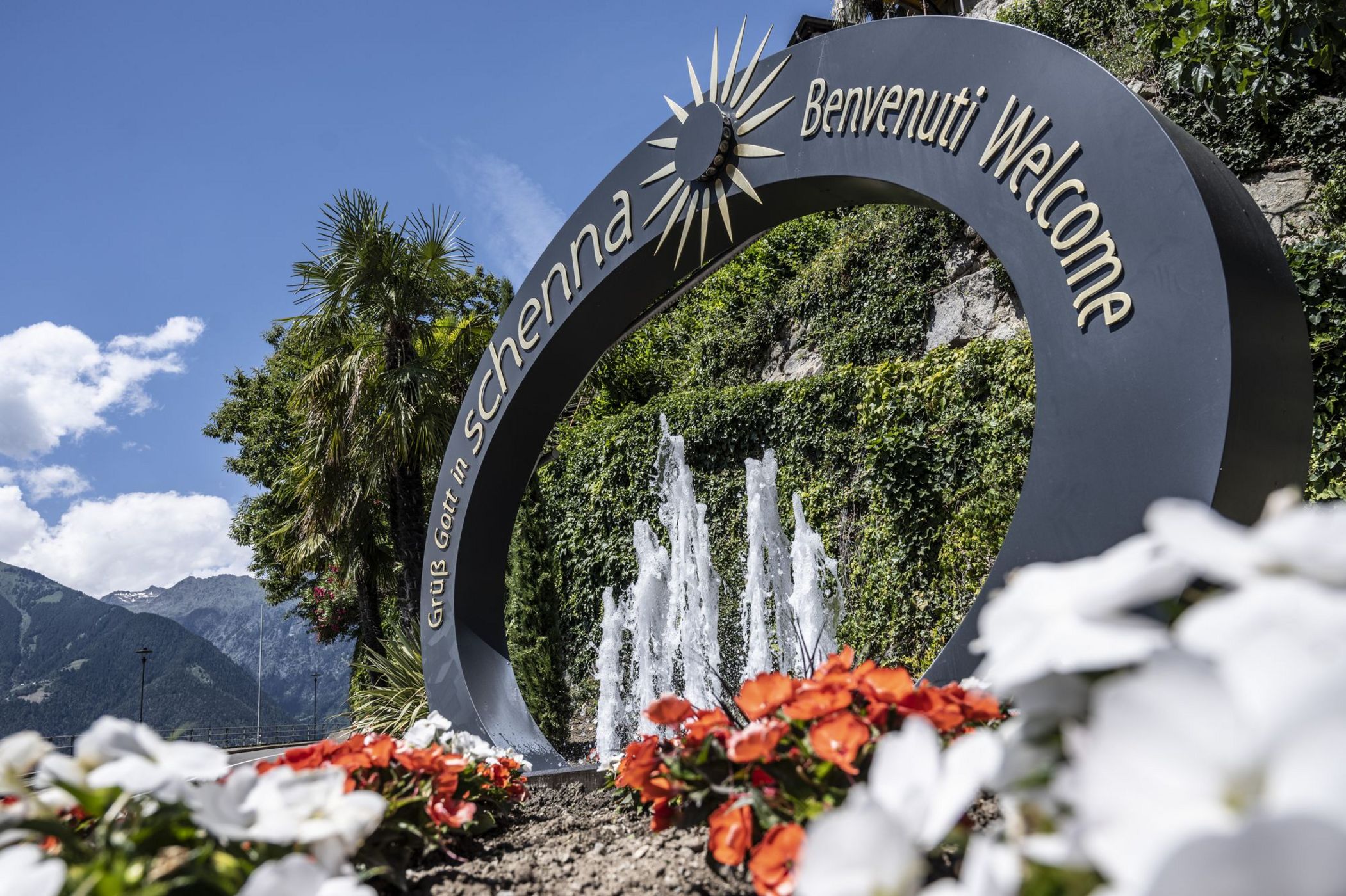 Fontana con arco e scritta di benvenuto in un giardino fiorito e montagne sullo sfondo